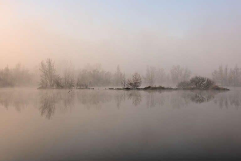 Photo d’un lac à l’aube avec un petit îlot au centre avec des arbres avec une lumière douce orangée pastelle