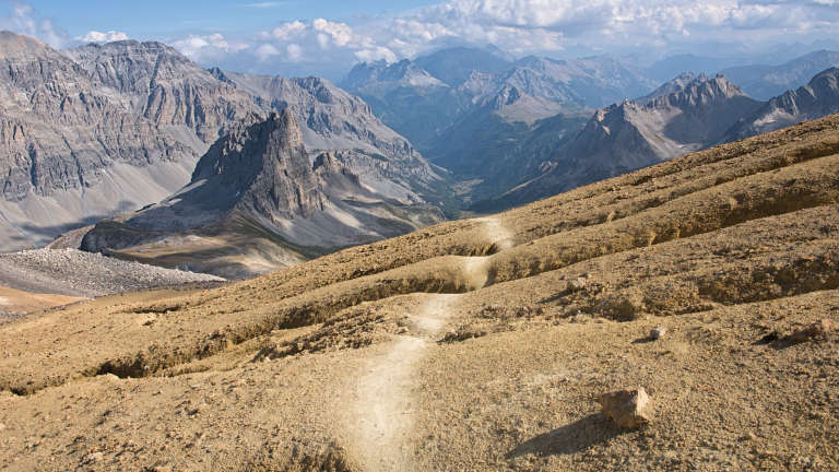 Photo d’un paysage montagneux avec un sol orange et des montagnes en arrière plan, un chemin laissé par les piétons et vélos se dirige vers le fond de l’image