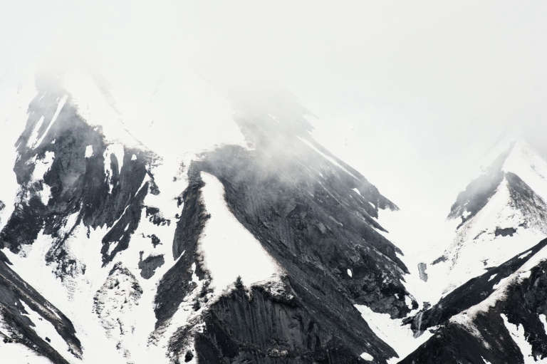 Photo en noir et blanc de montagnes noires avec des falaises avec de la neige et dans les nuages blancs opaques